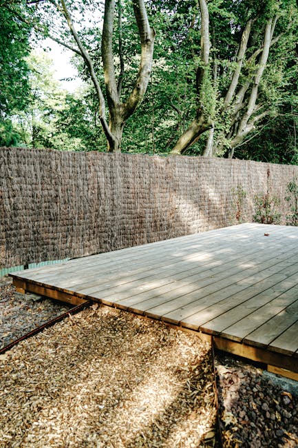 The image depicts a recently constructed wooden deck in a garden, with natural light filtering through the surrounding leafy trees. The deck is made of smooth, light-colored wooden planks laid out in
